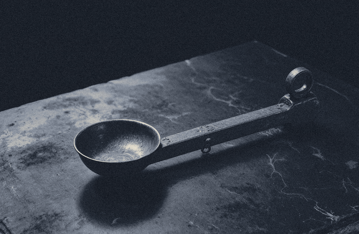 A small ancient bronze balance beam with one shallow pan hanging by fine cords, on dark slate, lit from one side.