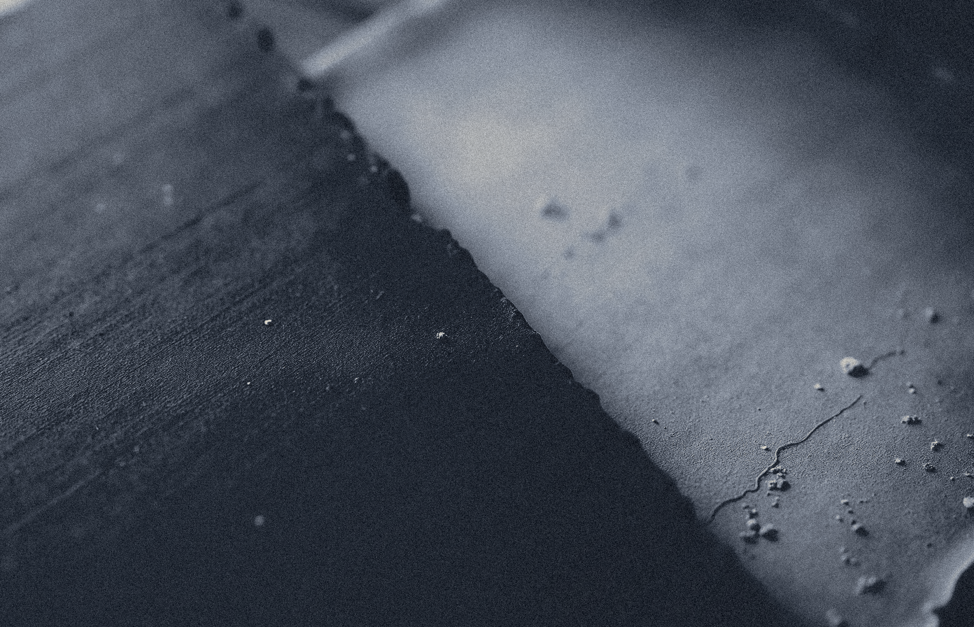 Macro photograph of the edge of a dark beeswax writing tablet beside the edge of a pale rough sheet, both blank, in raking light.